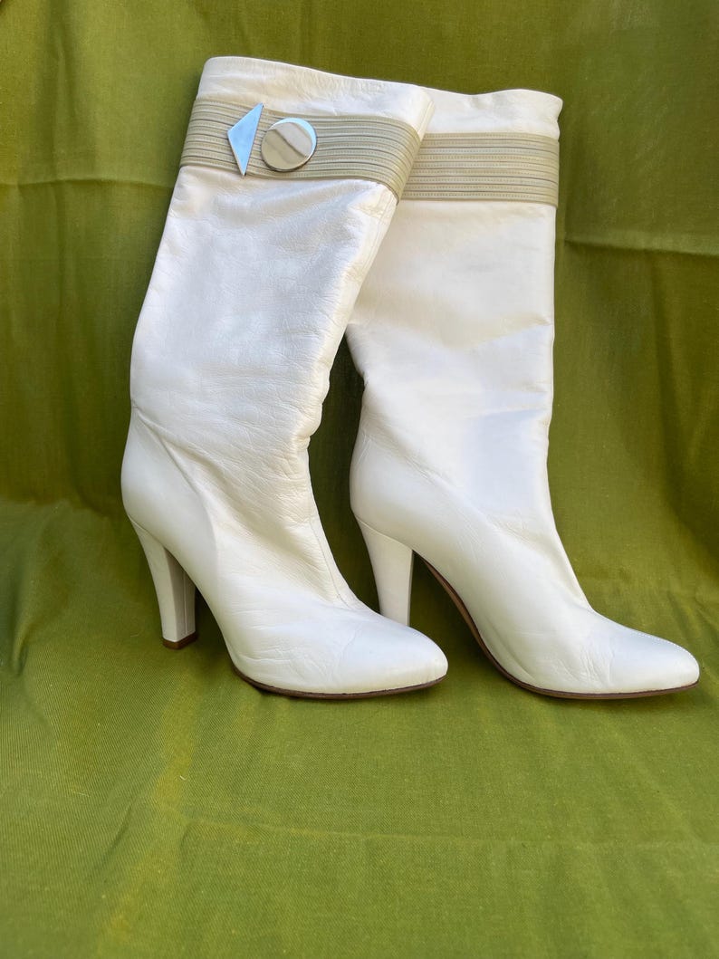 May include: A pair of white leather knee-high boots. The boots have a high heel and a decorative band around the top with a silver and beige accent. The boots are set against a green background.