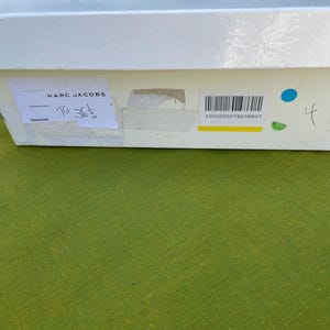 May include: A white rectangular box with a label that reads "MARC JACOBS". The label also includes the size "T6 36 1/2" and a barcode. The box is on a green surface.