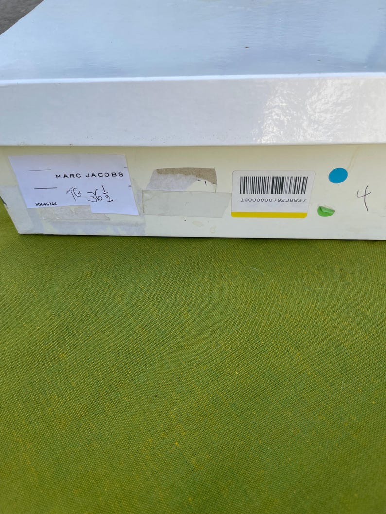 May include: A white rectangular box with a label that reads "MARC JACOBS". The label also includes the size "T6 36 1/2" and a barcode. The box is on a green surface.