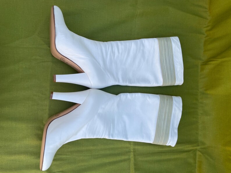 May include: A pair of white leather knee-high boots with a pointed toe and a stiletto heel. The boots have a beige sole and a beige elastic band detail at the top of the boot. The boots are set against a green background.