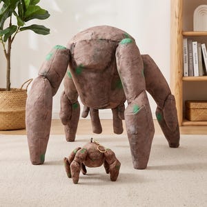 May include: A large, brown, rock-textured plush creature with multiple limbs and green moss-like patches, accompanied by a smaller, matching plush keychain version featuring a gold-toned metal ring attached to the top of its head.