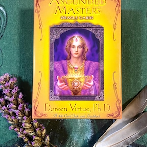 May include: A deck of "Ascended Masters Oracle Cards" by Doreen Virtue, Ph.D. The box is yellow with a central image of a woman in purple robes holding a golden chalice. The deck includes a 44-card deck and guidebook.