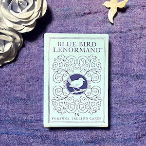 Może przedstawiać: Prostokątna talia kart do wróżenia Blue Bird Lenormand w jasnoniebieskim pudełku. Pudełko zawiera ciemnoniebieski tekst, centralną okrągłą ilustrację ptaka na gałęzi oraz skomplikowane wzory w kształcie zwojów na przedniej okładce.