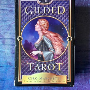 May include: A tarot card deck titled "The Gilded Tarot" with a woman in a blue dress and red hair on the front. The box is black with gold lettering and accents. The author's name, Ciro Marchetti, is also visible.