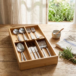 May include: A wooden cutlery organizer filled with stainless steel spoons, forks, and knives. The organizer sits on a wooden table, with a small ceramic bowl and a sprig of herbs nearby. The scene is lit by natural light.
