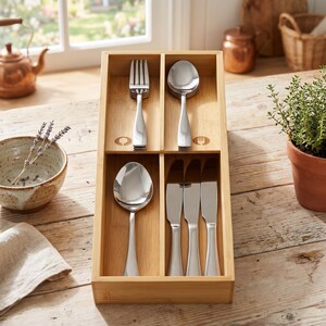May include: A wooden cutlery organizer with stainless steel silverware. The organizer has four compartments, each holding a different type of utensil: a fork, a spoon, and knives. The organizer is made of light-colored wood and sits on a wooden table.