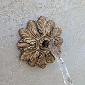 May include: A bronze-coloured water fountain spout with a floral design. Water flows from the centre of the spout. The spout is mounted on a textured, light-coloured wall. The design is reminiscent of classical architecture.
