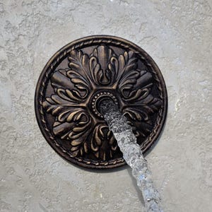 May include: A circular bronze-toned metal wall-mounted fountain fixture featuring an intricate embossed floral leaf pattern and a braided outer border, with a steady stream of clear water flowing from the center opening of the decorative plate.