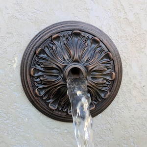 May include: A circular bronze-toned wall fountain spout featuring a raised, symmetrical floral relief pattern. Water flows continuously from the central opening of the medallion, creating a clear, vertical stream against the textured wall surface.