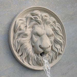 May include: A beige stone lion head fountain with water jetting from its mouth. The lion's face is intricately sculpted, set within a circular frame. The water stream is clear and flowing. The fountain is mounted on a textured wall.