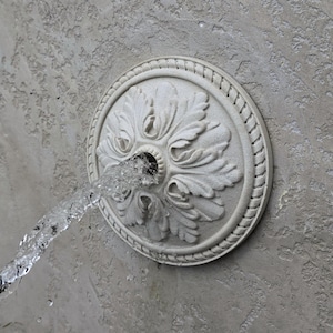 May include: A circular, off-white wall-mounted fountain spout featuring a central carved floral rosette design with a rope-textured border. Water flows continuously from the center of the medallion, which is mounted on a textured gray wall surface.