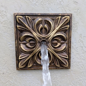 May include: A square wall-mounted fountain spout featuring a bronze-toned finish with dark antiqued highlights. The surface displays a symmetrical relief pattern of stylized acanthus leaves radiating from a central circular opening where water flows.