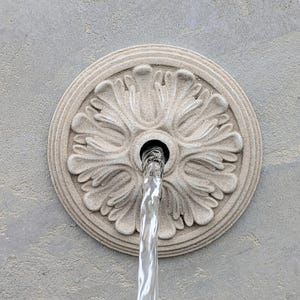 May include: A decorative, circular fountain element with a floral design. Water flows from a central opening, creating a visual contrast against the textured, light gray background. The fountain's intricate details are highlighted by the play of light and shadow.