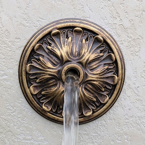May include: Ornate, bronze-colored fountain fixture with a floral design. Water flows from the center of the fixture. The fixture is circular and mounted on a textured, light-colored wall.