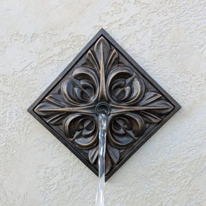 May include: Bronze-colored, diamond-shaped water fountain fixture with ornate, symmetrical floral design. Water flows from the center. The fixture is mounted on a textured, light-colored wall.