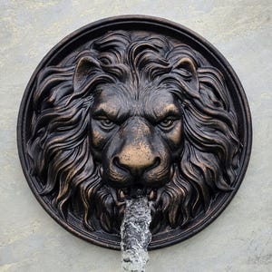 May include: A bronze-colored lion head fountain sculpture. The lion's face is detailed with a flowing mane and a stern expression. Water streams from the lion's mouth. The sculpture is circular and mounted on a light gray surface.