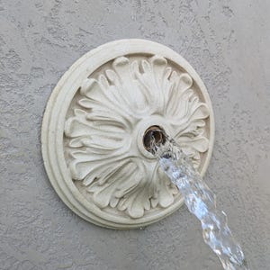 May include: A circular, off-white wall fountain spout featuring a detailed, carved floral or acanthus leaf relief pattern. A steady stream of clear water flows outward from the central metal opening, which is set against a textured, light gray wall.