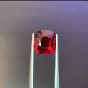 Könnte beinhalten: Ein facettierter, kissenförmiger, tiefroter Edelstein wird von einer silbernen Pinzette vor einem dunklen Hintergrund gehalten. Die vielen Facetten des Edelsteins reflektieren das Licht und erzeugen ein brillantes Farb- und Funkeln. Der Edelstein ist ein leuchtendes Rot.