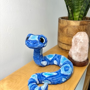 Könnte beinhalten: Ein blaues Plüsch-Schlangenspielzeug mit einem gemusterten Design in Blau- und Weißtönen. Die Schlange hat große schwarze Augen und liegt auf einem Holzregal. Es ist ein skurriles Dekorationsstück.