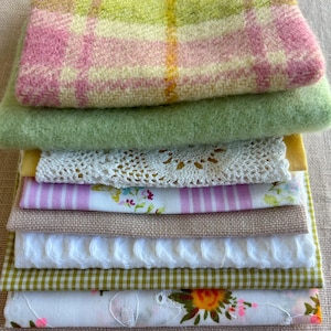 May include: A stack of folded fabrics including a pink and lime green plaid wool, a solid lime green felt, a white lace doily, a pink and white floral print, a tan linen, a white cable knit, a green gingham, and a white floral embroidered cotton.