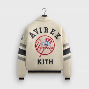 May include: An off-white varsity jacket featuring navy blue and grey striped sleeves. The back displays an embroidered Avirex logo, a New York Yankees baseball graphic with a top hat, and the word Kith in navy blue lettering on the lower back.
