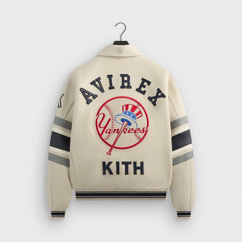 May include: An off-white varsity jacket featuring navy blue and grey striped sleeves. The back displays an embroidered Avirex logo, a New York Yankees baseball graphic with a top hat, and the word Kith in navy blue lettering on the lower back.