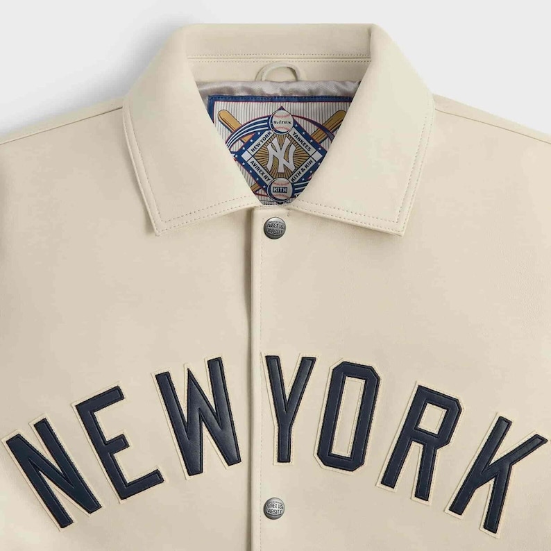 May include: A cream-colored jacket featuring dark navy blue lettering that spells NEW YORK across the chest. The collar reveals an interior label with a baseball-themed graphic containing the text New York Yankees, Avirex NY, and Kith & Kin.
