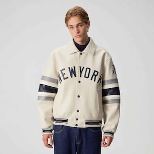 May include: A cream-colored varsity jacket featuring a dark navy blue arched NEW YORK text across the chest. The sleeves have navy blue and grey horizontal stripes, and the jacket includes a snap-button front closure with ribbed cuffs and hem.