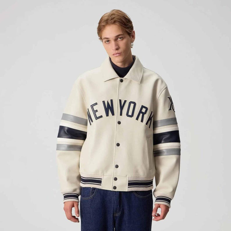 May include: A cream-colored varsity jacket featuring a dark navy blue arched NEW YORK text across the chest. The sleeves have navy blue and grey horizontal stripes, and the jacket includes a snap-button front closure with ribbed cuffs and hem.