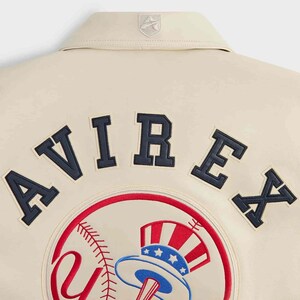 May include: A cream-colored jacket featuring the word AVIREX in dark navy blue block letters across the upper back. Below the text is a circular patch depicting a baseball with red stitching and a red, white, and blue top hat graphic.