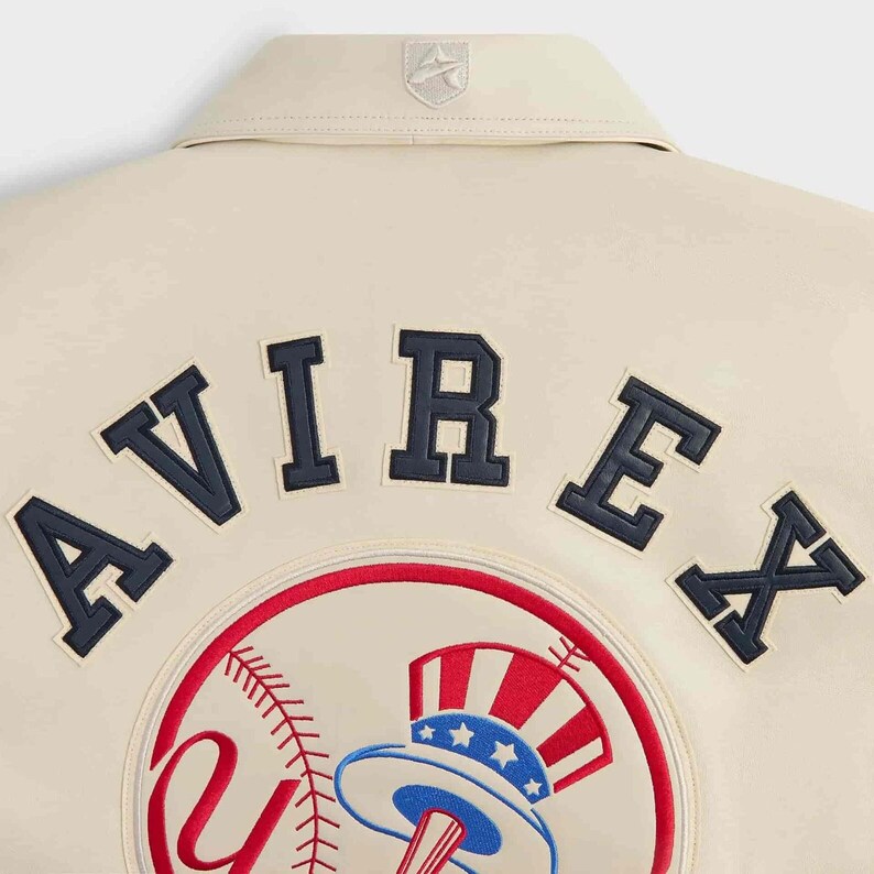 May include: A cream-colored jacket featuring the word AVIREX in dark navy blue block letters across the upper back. Below the text is a circular patch depicting a baseball with red stitching and a red, white, and blue top hat graphic.