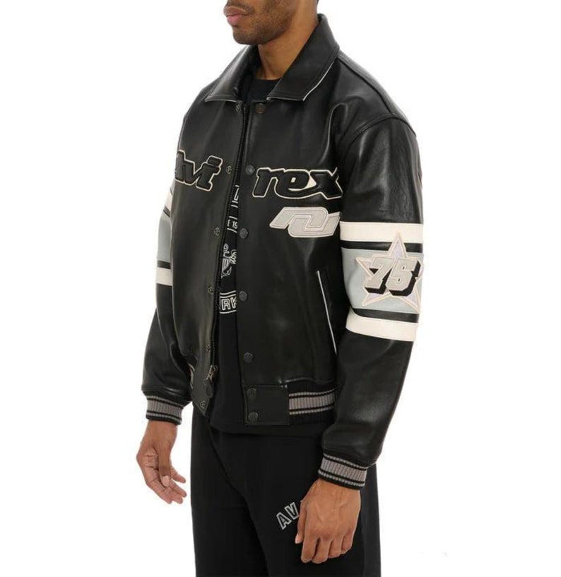 May include: Black leather jacket with white and gray accents. The jacket features the word "M-rex" across the chest and "78" on the sleeve. The jacket has a ribbed collar, cuffs, and hem.