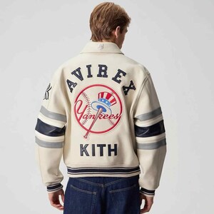 May include: An off-white varsity jacket featuring navy blue and grey striped sleeves. The back displays the text AVIREX and KITH in navy blue lettering, with a central embroidered New York Yankees logo featuring a baseball and a top hat.