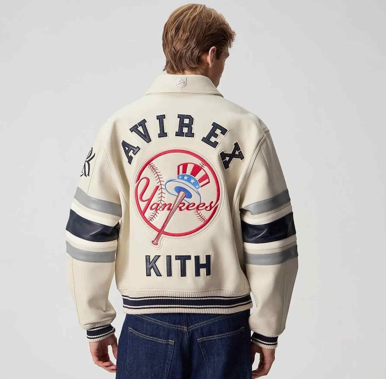 May include: An off-white varsity jacket featuring navy blue and grey striped sleeves. The back displays the text AVIREX and KITH in navy blue lettering, with a central embroidered New York Yankees logo featuring a baseball and a top hat.