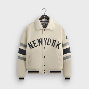 May include: A cream-colored varsity jacket featuring the text NEW YORK in dark navy blue lettering across the chest. The sleeves have navy and grey stripes, and the jacket includes a button-front closure, a collar, and a Kith logo on the left sleeve.