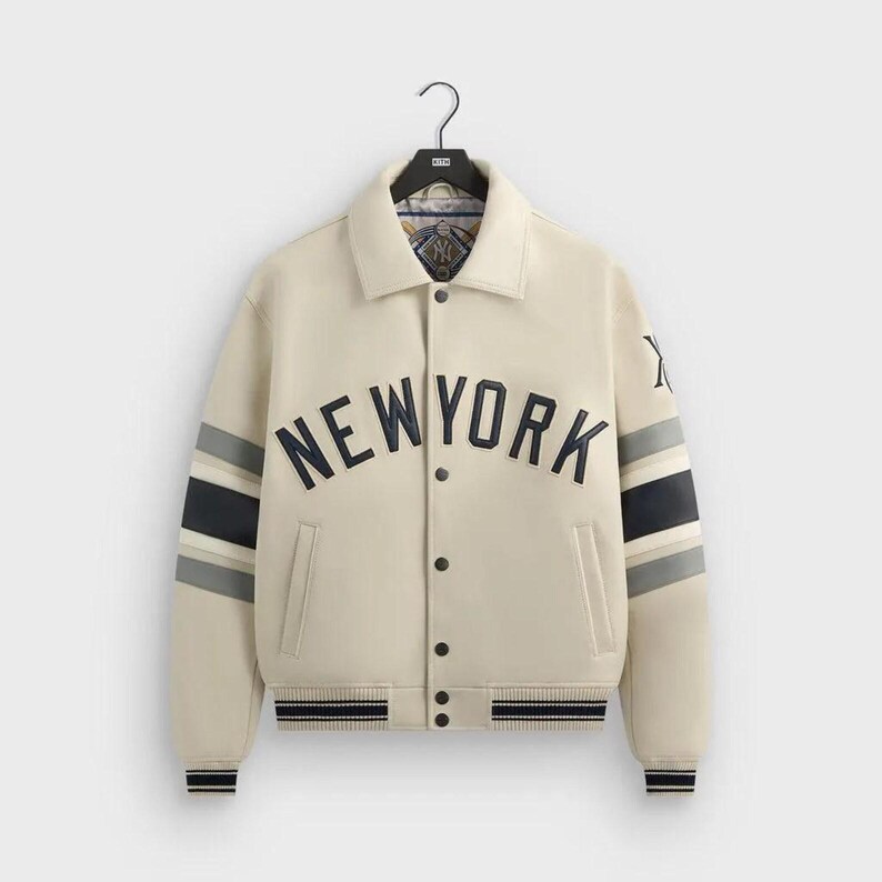 May include: A cream-colored varsity jacket featuring the text NEW YORK in dark navy blue lettering across the chest. The sleeves have navy and grey stripes, and the jacket includes a button-front closure, a collar, and a Kith logo on the left sleeve.