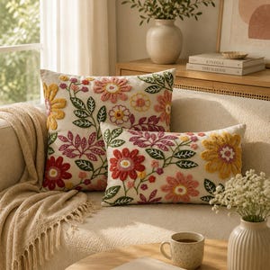 Può includere: Due cuscini decorativi con ricami floreali rossi, gialli e rosa su sfondo crema. I cuscini sono su un divano beige, con una coperta con frange e un tavolino con un libro e una tazza. Atmosfera accogliente.