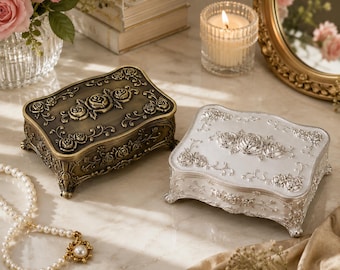 May include: Two rectangular jewelry boxes with ornate floral relief patterns. One box features a dark antique bronze finish, while the other has a bright silver finish. Both boxes stand on four decorative feet and have matching rose motifs.