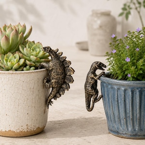 May include: Two bronze dinosaur pot hangers, a Stegosaurus and a Tyrannosaurus Rex, cling to the edges of ceramic pots. One pot holds a succulent, the other a flowering plant. The pots are in neutral tones, creating a natural, decorative theme.