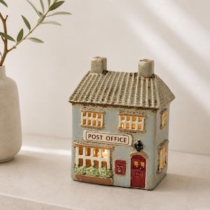 Puede incluir: Una figura de cerámica de oficina de correos con una fachada azul claro, un tejado gris y una puerta roja. Las ventanas brillan con luz cálida, y el letrero dice "POST OFFICE". Un artículo decorativo para la decoración del hogar.