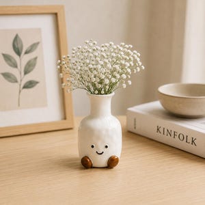 May include: A white ceramic vase with a smiling face and brown feet holds a bouquet of white flowers. The vase is on a wooden surface, next to a framed botanical print and a book titled "KINFOLK". The vase is approximately 13 cm tall.