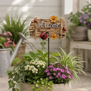 Op de afbeelding: Een decoratief tuinbord met het woord "Welcome" in witte letters. Het bord is gemaakt van verweerd hout en heeft lieveheersbeestjes en gele bloemen. Het staat in een pot met diverse groene planten en roze en witte bloemen.