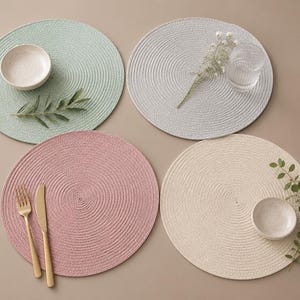 Puede incluir: Cuatro manteles individuales redondos trenzados en verde salvia, gris claro, rosa empolvado y crema. Los manteles presentan una textura en espiral concéntrica. Incluye dos cuencos de cerámica, un vaso transparente, cubiertos dorados y ramitas verdes.