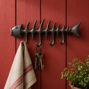 May include: A cast iron fish skeleton wall hook with multiple hooks for hanging items. The dark metal hook is mounted on a red wooden wall, with a towel and keys hanging from the hooks. A small potted plant is in the lower right corner.