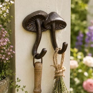 May include: A dark brown cast iron wall hook featuring two mushroom-shaped designs. Each mushroom cap serves as a mounting point with a visible screw, while the curved stems extend downward to form functional hooks for hanging various items.