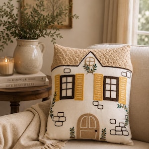May include: A cushion with a house design. The pillow has a white background with embroidered details, including brown window frames, yellow shutters, and a tan thatched roof. Green embroidered vines and brick accents add detail.