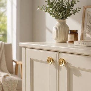 May include: Cream-coloured cabinet with gold flower-shaped knobs. A white vase with greenery, a brown jar, and a framed picture sit on top. The cabinet doors have a recessed panel design.