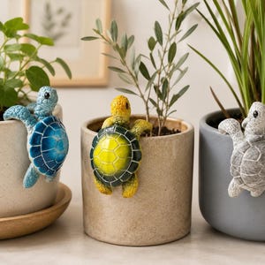 Op de afbeelding: Drie keramische plantenpotten met decoratieve schildpadfiguren. De schildpadden zijn blauw, geel en grijs en lijken over de randen van de potten te klimmen. De potten bevatten verschillende groene planten, wat een grillige en decoratieve weergave creëert.