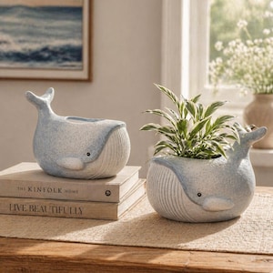 May include: Two light blue whale-shaped planters. One planter is empty, and the other contains a green and white plant. The planters have a textured finish and are sitting on a wooden table. The planters are next to a stack of books.