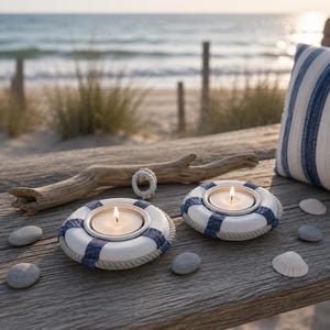 May include: Two white and blue nautical-themed candle holders shaped like life preservers, each with a lit candle. The holders are on a weathered wooden surface, with a driftwood piece, pebbles, and a striped pillow in the background.